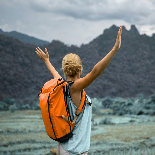 Outdoor adventure lifestyle image showing a hiker with orange backpack raising arms in front of mountain landscape